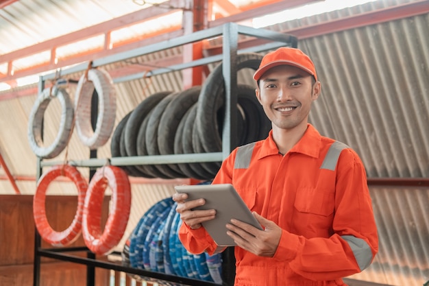 Aziatische lachende monteur in wearpack met behulp van een stootkussen terwijl in de werkplaats met een bandenrek erachter