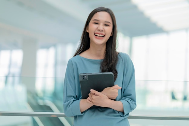 Aziatische lachende vrolijke vrouwelijke digitale nomade hand houden tablet-apparaat kijken camera portret shothappiness lachende Azië vrouw staande in kantoor college gang met positieve lachende houding