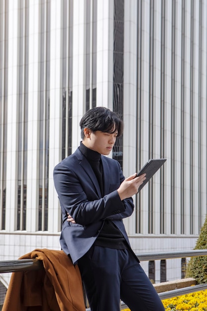 Aziatische man buiten het zakencentrum die op zijn mobiele telefoon praat terwijl hij naar zijn tablet kijkt