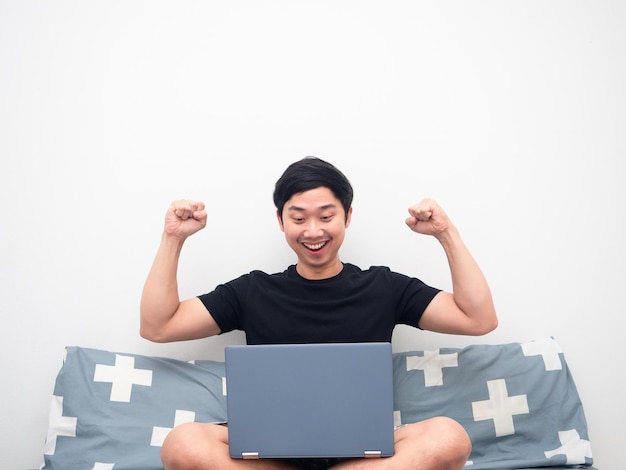 Aziatische man die naar laptop kijkt en zijn vuist laat zien, voelt zich gelukkig in de slaapkamer