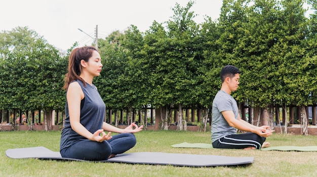 Aziatische man en vrouw beoefenen van yoga buitenshuis in mediteren pose zittend op groen gras. Jong stel dat zich in de natuur uitstrekt in een veldtuinpark. Meditatie, oefen gezondheidszorgconcept