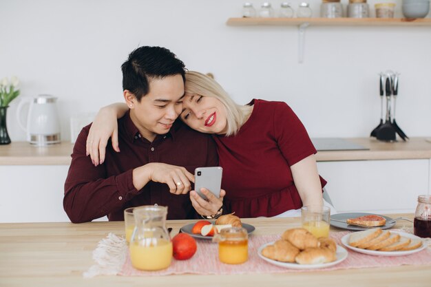 Aziatische man en zijn blanke blonde vrouw ontbijten. ze brengen samen tijd door in de keuken en kijken video aan de telefoon.