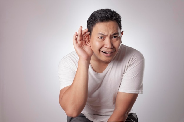 Aziatische man houdt zijn hand op het oor probeert om aandachtig te luisteren naar het gesprek nieuwsgierige uitdrukking