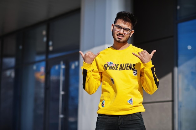 Aziatische man in brillen en geel sweatshirt poseren tegen een nieuw modern gebouw en tonen een cool teken met de vingers