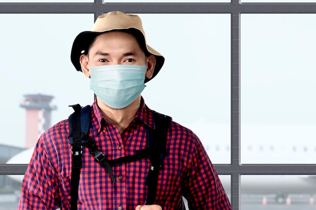 Aziatische man in het gezichtsmasker met hoed en een rugzak op de luchthaventerminal. Reizen in het nieuwe normaal