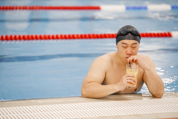 Aziatische man in hoed en bril die een sinaasappelsap drinkt in het zwembad