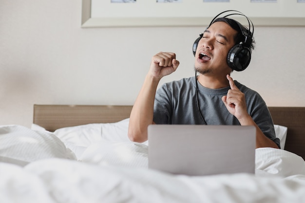 Aziatische man in hoofdtelefoon luistert naar een liedje van laptop terwijl hij op bed zingt