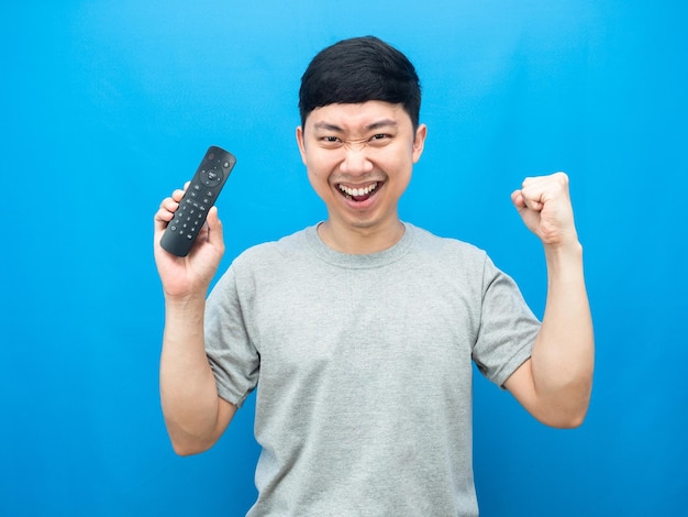 Aziatische man met afstandsbediening televisie toont vuist blije emotie blauwe achtergrond