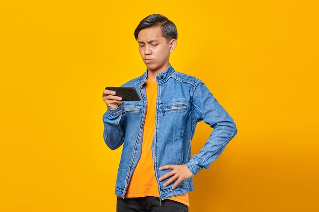 Aziatische man met behulp van mobiele telefoon speelspel met boos gezicht over gele achtergrond