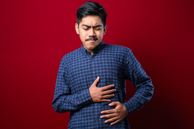 Aziatische man met een casual shirt diarree gezondheidsproblemen houdt zijn buik op een rode achtergrond
