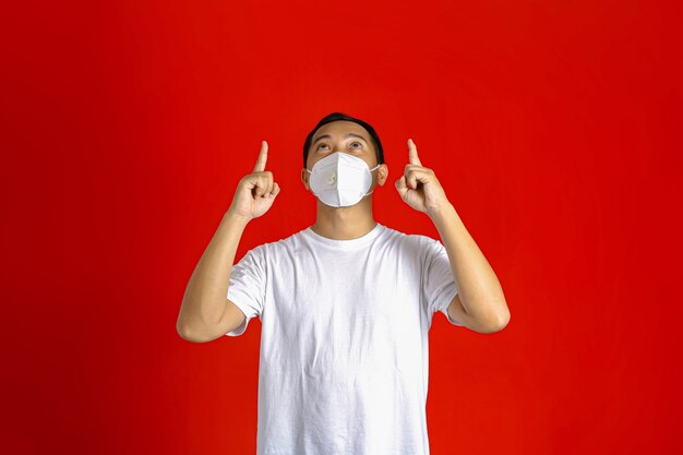 Aziatische man met een medisch masker die met beide handen omhoog wijst terwijl hij omhoog kijkt op een rode achtergrond