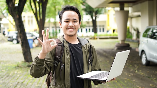 Aziatische man met een tas terwijl hij een laptop vasthoudt met ok gebaar