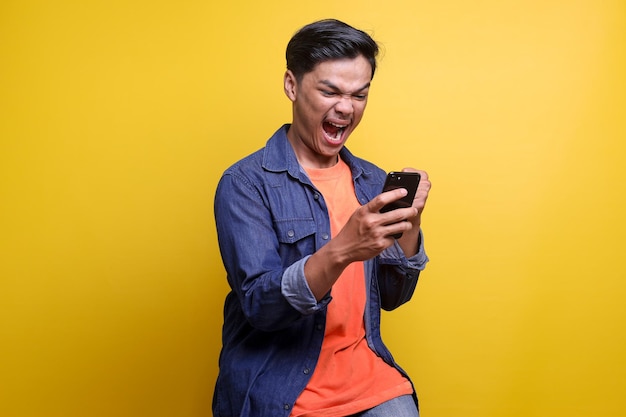 Aziatische man met een uitdrukking van woede als hij naar zijn mobiele telefoon kijkt tegen een gele achtergrond een advertentie