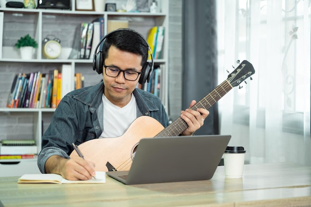 Aziatische man met koptelefoon speelt gitaar en zingt een lied en leert online met laptop en notitieboekje en componeert het schrijven van liedjes, neemt geluid op en gebruikt computeropname muziekprogramma Muzikant concept