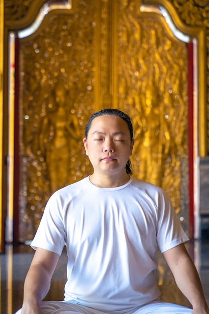 Aziatische man met lang haar ontspant meditatie met een geheel wit kostuum, zittend voor het gouden behang van Buddist in de tempel, Thailand.