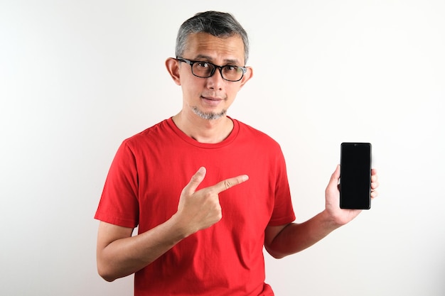 Aziatische man met rode t-shirt op zoek naar camera met smartphone leeg scherm witte achtergrond