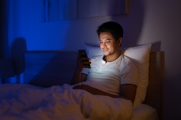 Aziatische man typt online chat met een vriend of vriendin 's nachts op een bed in een slaapkamer die uit het licht is.