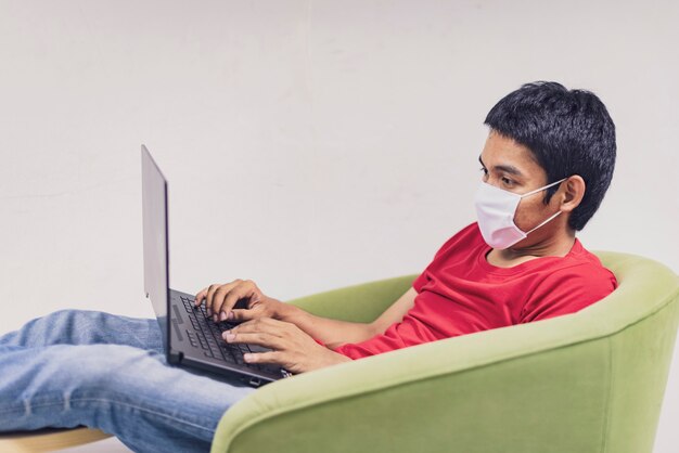 Aziatische man werkt vanuit huis tijdens Coronavirus of Covid-19. Draag een gezichtsmasker ter bescherming tegen coronavirus, werk thuis en gebruik een laptop. Werk vanuit huis, blijf thuis. Sociale afstand.