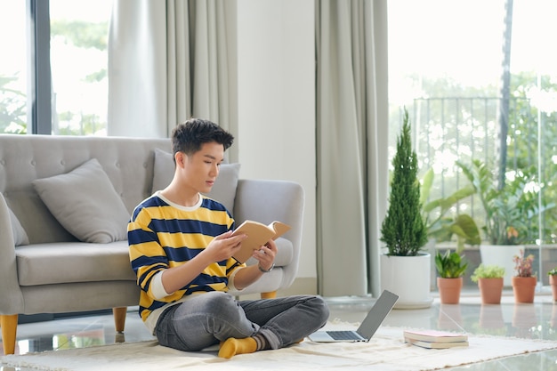 Aziatische man zit op de grond met een boek en laptop