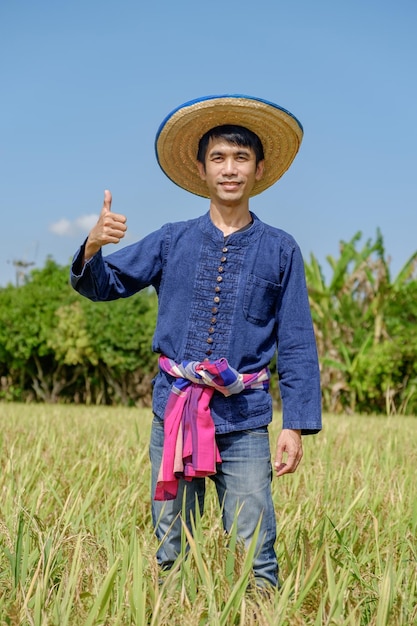Aziatische mannelijke boer met een blauw shirt met een hoed op en duim omhoog met een lachend gezicht naar het rijstveld