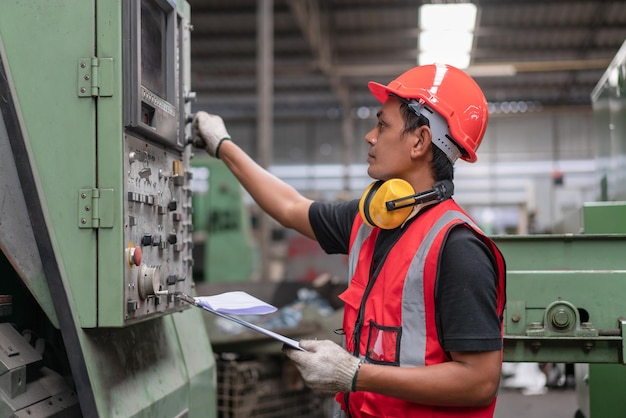 Aziatische mannelijke ingenieur in rood veiligheidsvest en helm die oude CNC-machine in de fabriek controleert en repareert