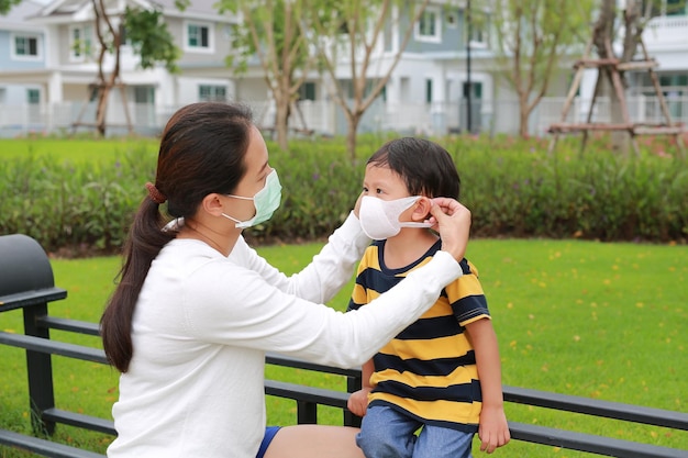 Aziatische moeder die beschermend gezichtsmasker voor babyjongen in de openbare tuin draagt tijdens coronavirus en griepuitbarsting