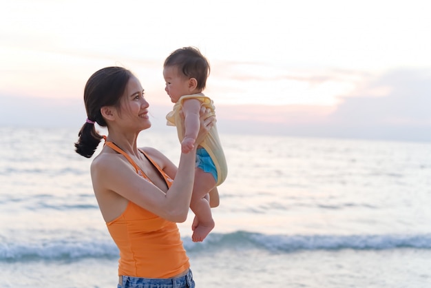 Aziatische moeder die zich op het strand bevindt dat haar baby in twee wapens houdt die het jonge geitje opheffen en haar kind bekijken.