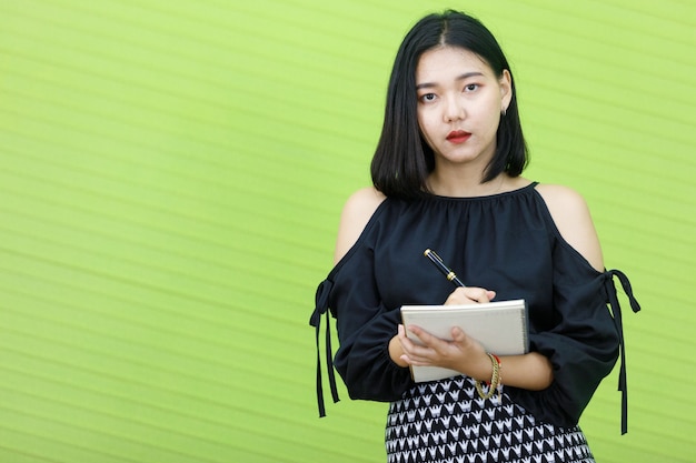 Aziatische mollige vrouw die zwarte kleding draagt. Zakenvrouw met handboek en pen op groene achtergrond. Concept mooie dame die zelfverzekerd werkt.