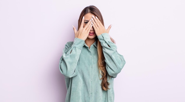 Aziatische mooie vrouw die haar gezicht bedekt met handen, tussen de vingers gluurt met een verbaasde uitdrukking en naar de zijkant kijkt