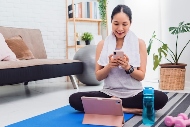 Aziatische mooie vrouw oefenen thuis en kijken naar trainingsvideo op smartphone en tablet.