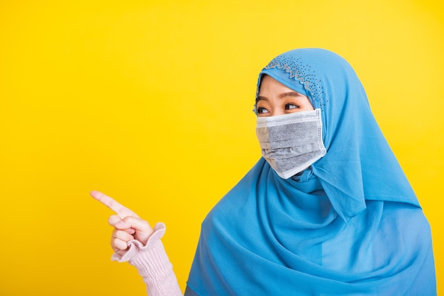 Aziatische moslim Arabier, Portret van gelukkige mooie jonge vrouw religieuze slijtage sluier hijab en gezichtsmasker beschermend om coronavirus te voorkomen zij wijzende vinger zijwaarts weg ruimte, geïsoleerd op gele achtergrond