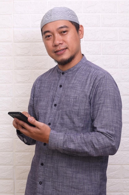 Aziatische moslim man die lacht blij wanneer hij zijn mobiele telefoon gebruikt terwijl hij naar de camera kijkt