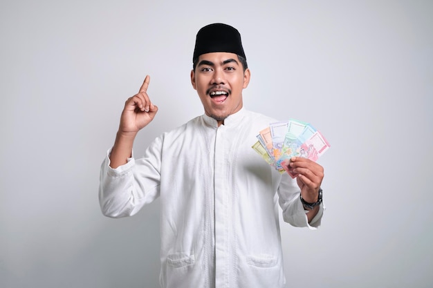 Aziatische moslim man die rupiah papiergeld vasthoudt terwijl hij met zijn vinger omhoog wijst en glimlacht