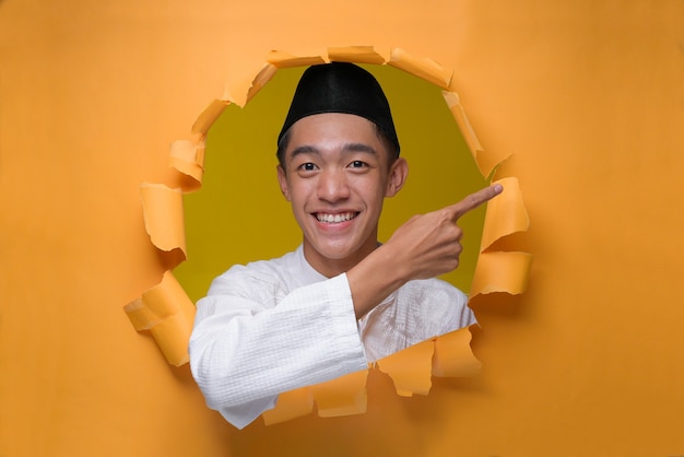 Aziatische moslim tiener man glimlachend poses door gescheurd geel papier gat, moslim doek dragen met schedeldak, een kopie ruimte wijzend om iets te presenteren.