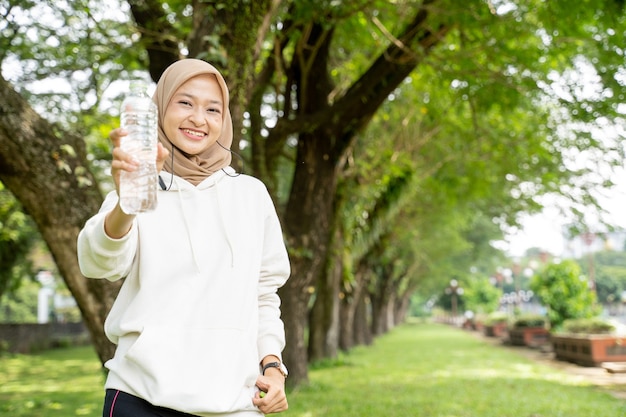 Aziatische moslimvrouw die met hoofddoek een fles water drinkt tijdens het sporten buiten