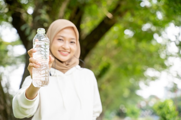 Aziatische moslimvrouw die met hoofddoek een fles water drinkt tijdens het sporten buiten