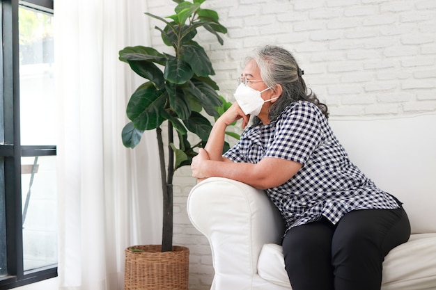 Aziatische oudere vrouwen met maskers ze zat eenzaam op de bank in het huis. thuis voor jezelf zorgen tijdens de covid-19 pandemie. zelfzorg om besmetting met coronavirus te voorkomen, thuisisolatie