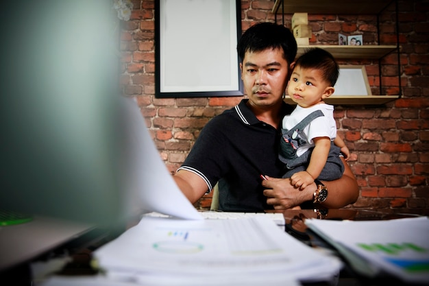 Aziatische papa houdt zijn pasgeboren zoontje vast en werkt thuis met laptop