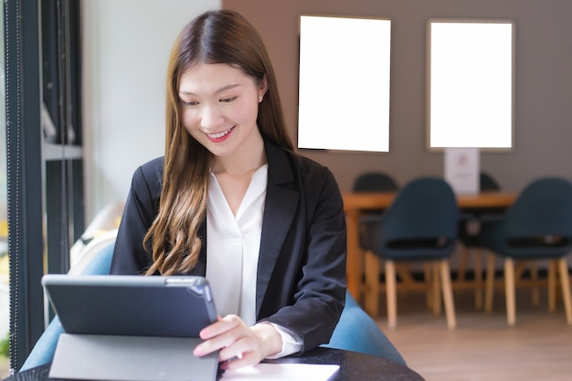 Aziatische professionele werkende vrouw in een zwart pak werkt aan een tablet op de tafel die vrolijk glimlacht op kantoor en thuis werkt.