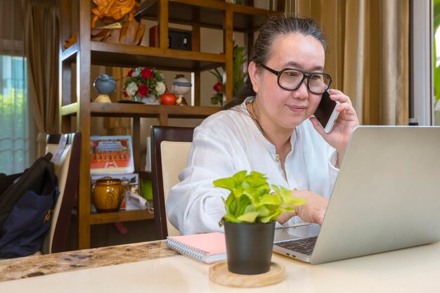 Aziatische rijpe vrouw die op smartphone praat en laptopcomputer gebruikt terwijl ze thuis werkt