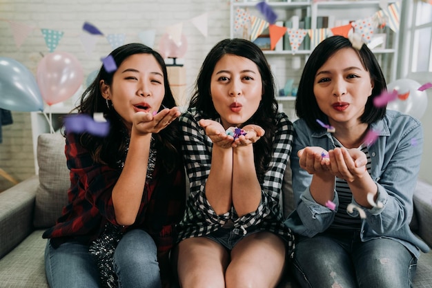 Aziatische schattige dames worden geconfronteerd met camera die vrolijk kleurrijke confetti blaast. jonge meisjes zitten ontspannen op de bank in ingerichte woonkamer thuis. huisfeest vol ballonnen vrienden die plezier hebben met vieren.