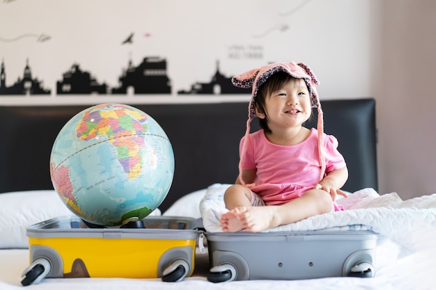 Aziatische schattige kleine baby meisje draagt hoed zittend op reistas met glimlach gevoel grappig en lachen op bed in de slaapkamer met wereldbol aan de andere kant van koffer tas.