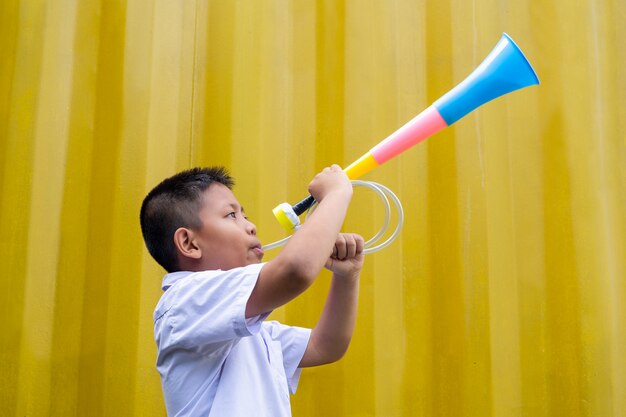 Aziatische schooljongen die een kleurrijke hoorn op gele achtergrond blaast.