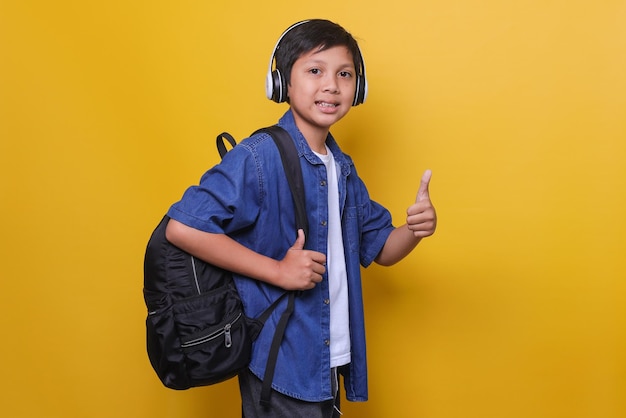 Aziatische schooljongen in casual denimstijl met koptelefoon en rugzak die glimlach en duim omhoog isolat