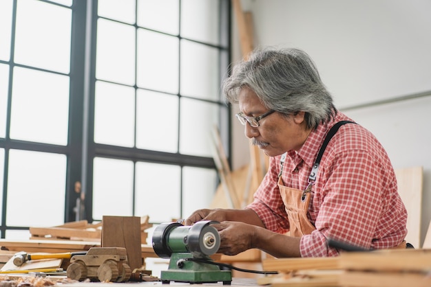 Aziatische senior timmerman man met behulp van kleine slijpmachine maken houten auto op workshop