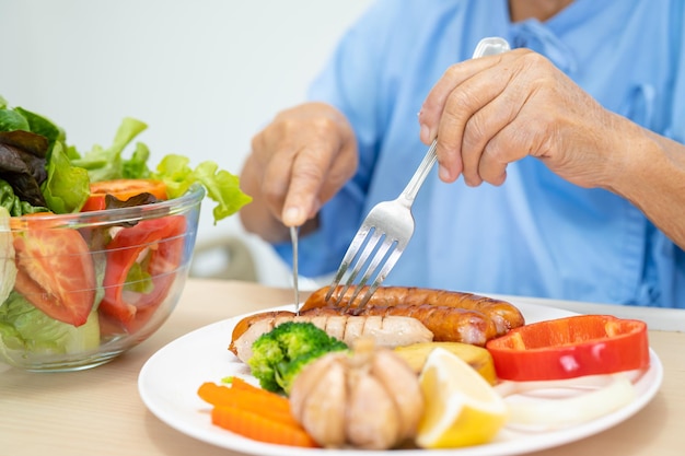 Aziatische senior vrouw patiënt eet zalm steak ontbijt met plantaardig gezond voedsel zittend en hongerig op bed in het ziekenhuis