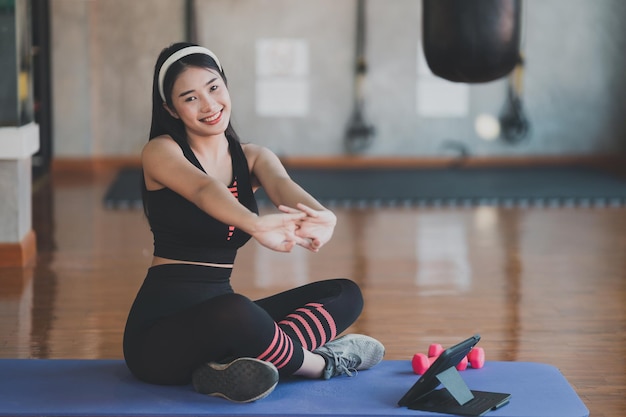 Aziatische sportvrouw zittend op yogamat en rekoefeningen doen met haltertraining online en kijken naar videostreaming op tablet binnen in de fitnessruimte Online fitness training gym concept