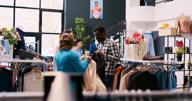 Aziatische stijlvolle werknemer helpt paar met formele outfit, bespreekt merchandise-stof in moderne boetiek. Afro-Amerikaanse klanten winkelen voor modieuze kleding in kledingwinkel