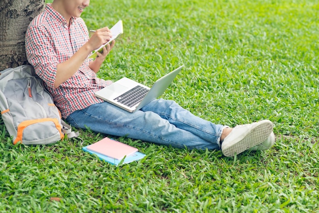 Aziatische student die composities schrijft in voorbeeldenboeken die op het gazon van het park zitten