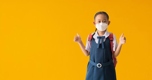 Aziatische student meisje draagt medisch gezichtsmasker met duimen omhoog voor goed geïsoleerd op gele achtergrond met uitknippaden voor ontwerp werk lege vrije ruimte